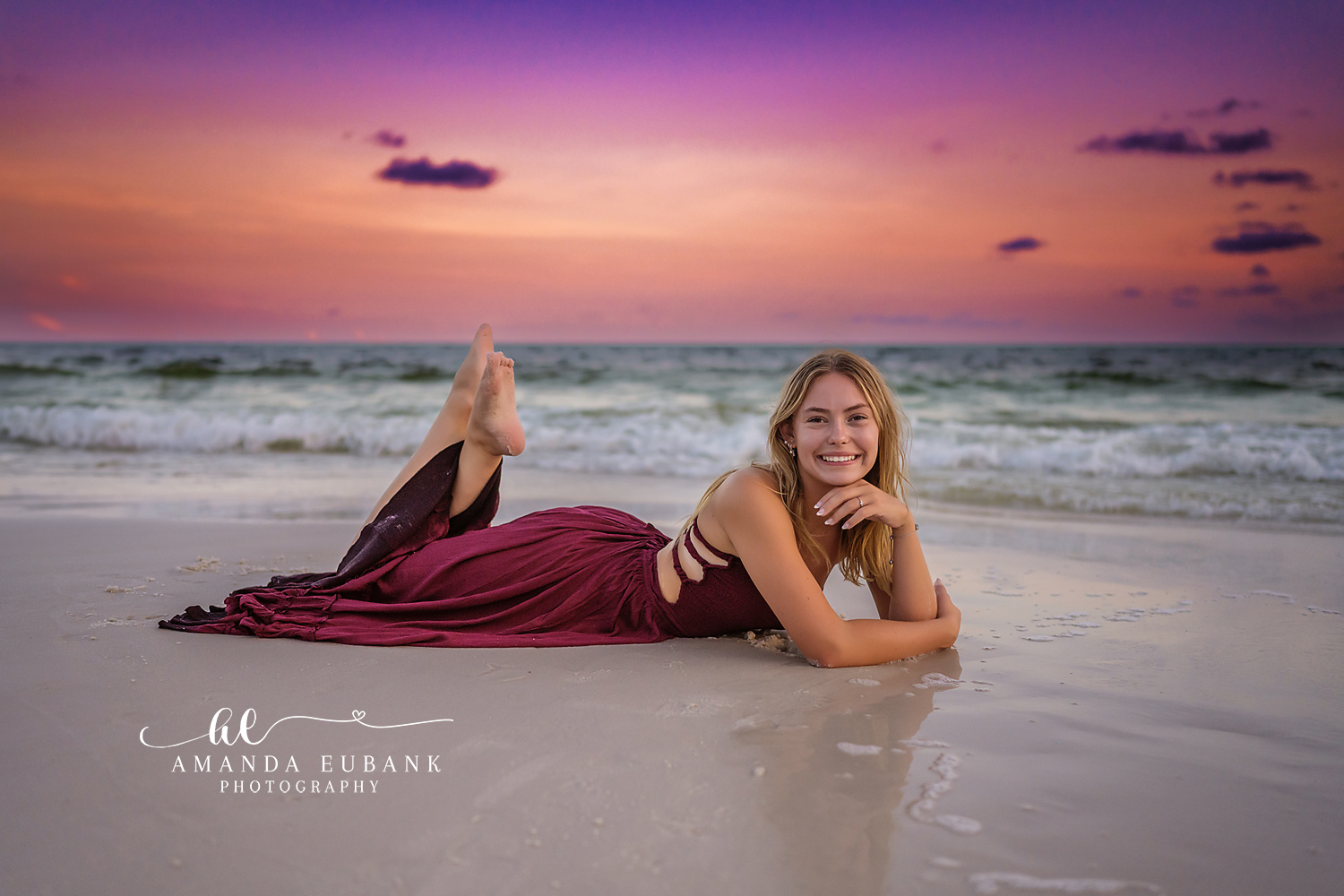 Senior girl in a burgundy gown lying on the wet sand propped on her elbows at the shoreline during a pink sunset, photographed by a Santa Rosa Beach senior photographer