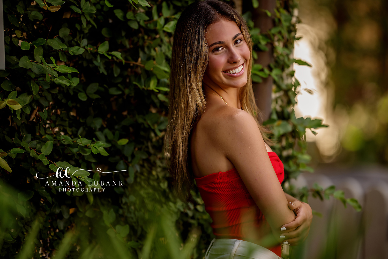 Senior girl in a red strapless top posed in profile among lush green foliage at golden hour, photographed by a Santa Rosa Beach senior photographer