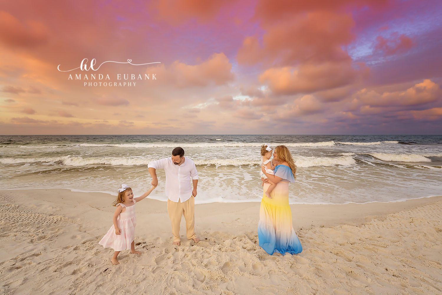 Family of four walking on the beach at sunset, the father holding a young daughter’s hand and the mother carrying a baby, photographed by a Santa Rosa Beach family photographer