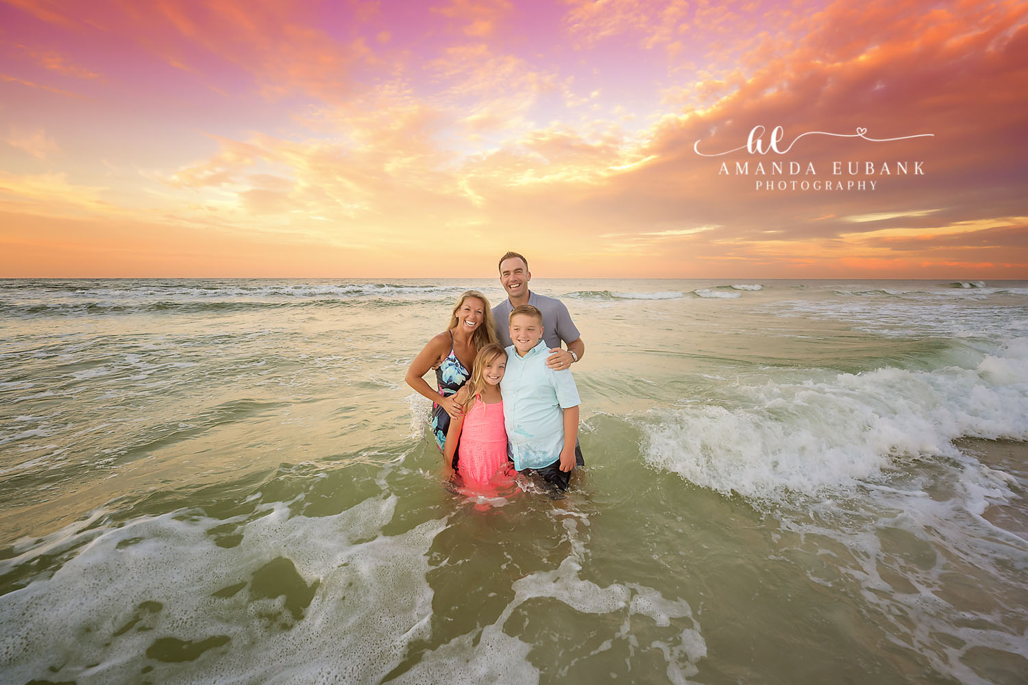Family of four standing together in the surf at sunset as a wave breaks around them, photographed by a Santa Rosa Beach family photographer