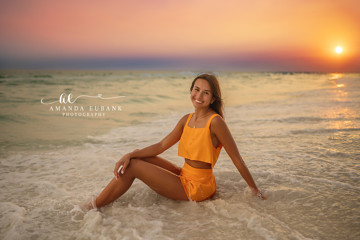 Senior girl in a bright orange two-piece set sitting in the shallow surf at golden hour, photographed by a Santa Rosa Beach senior photographer