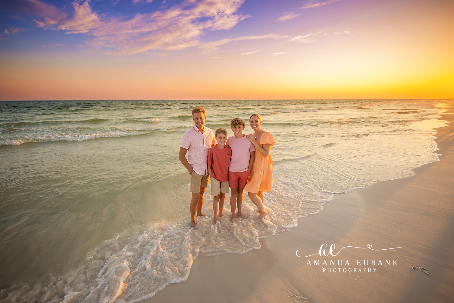 Family of four in coordinated coral outfits standing together at the shoreline at sunset, the parents with their two sons, photographed by a Santa Rosa Beach family photographer