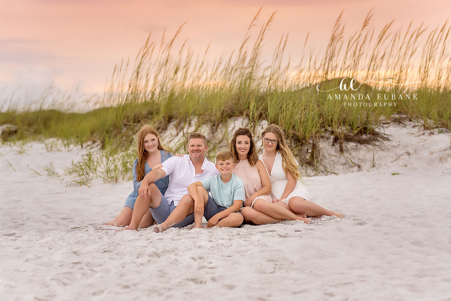 Family of five sitting together on the sand among the dune grasses in coordinated neutral and blue outfits, photographed by a Santa Rosa Beach family photographer