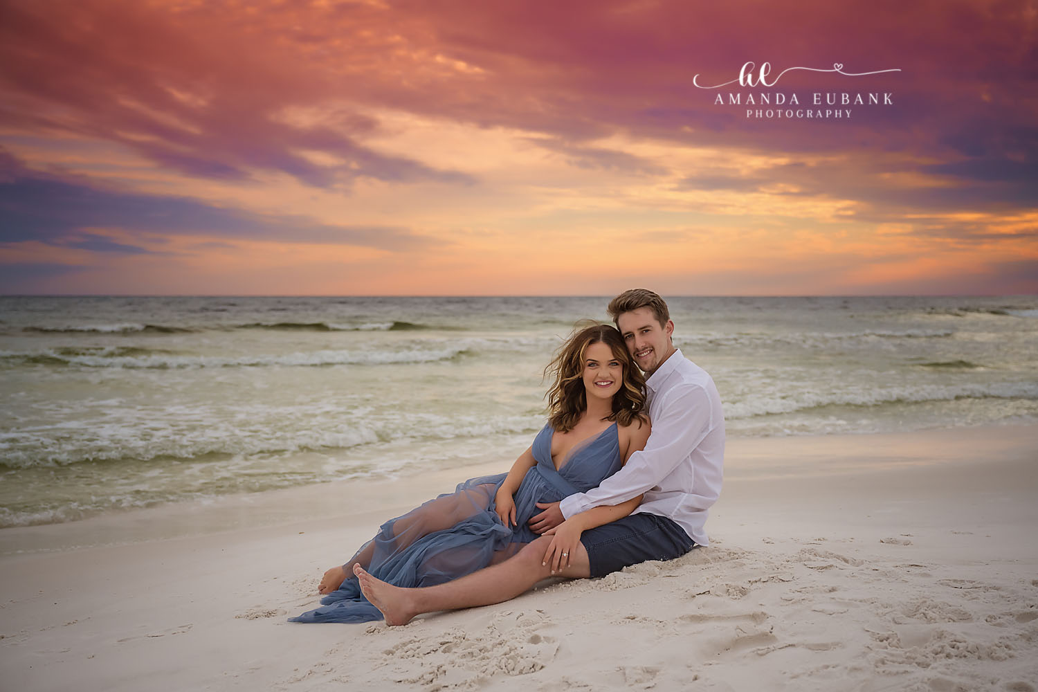 Expecting couple sitting on the sand at sunset, the woman in a flowing dusty blue maternity gown as both cradle her belly, photographed by a Santa Rosa Beach maternity photographer