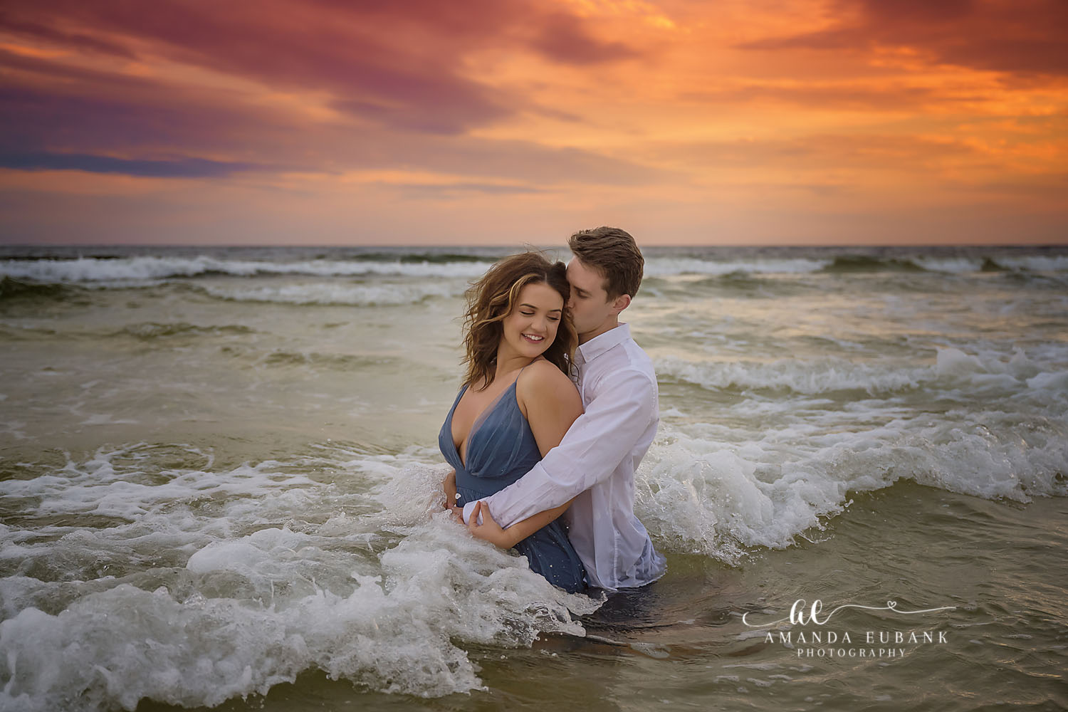 Couple embracing in the breaking surf at sunset, the man kissing the woman’s head as she wears a blue dress, photographed by a Santa Rosa Beach engagement photographer