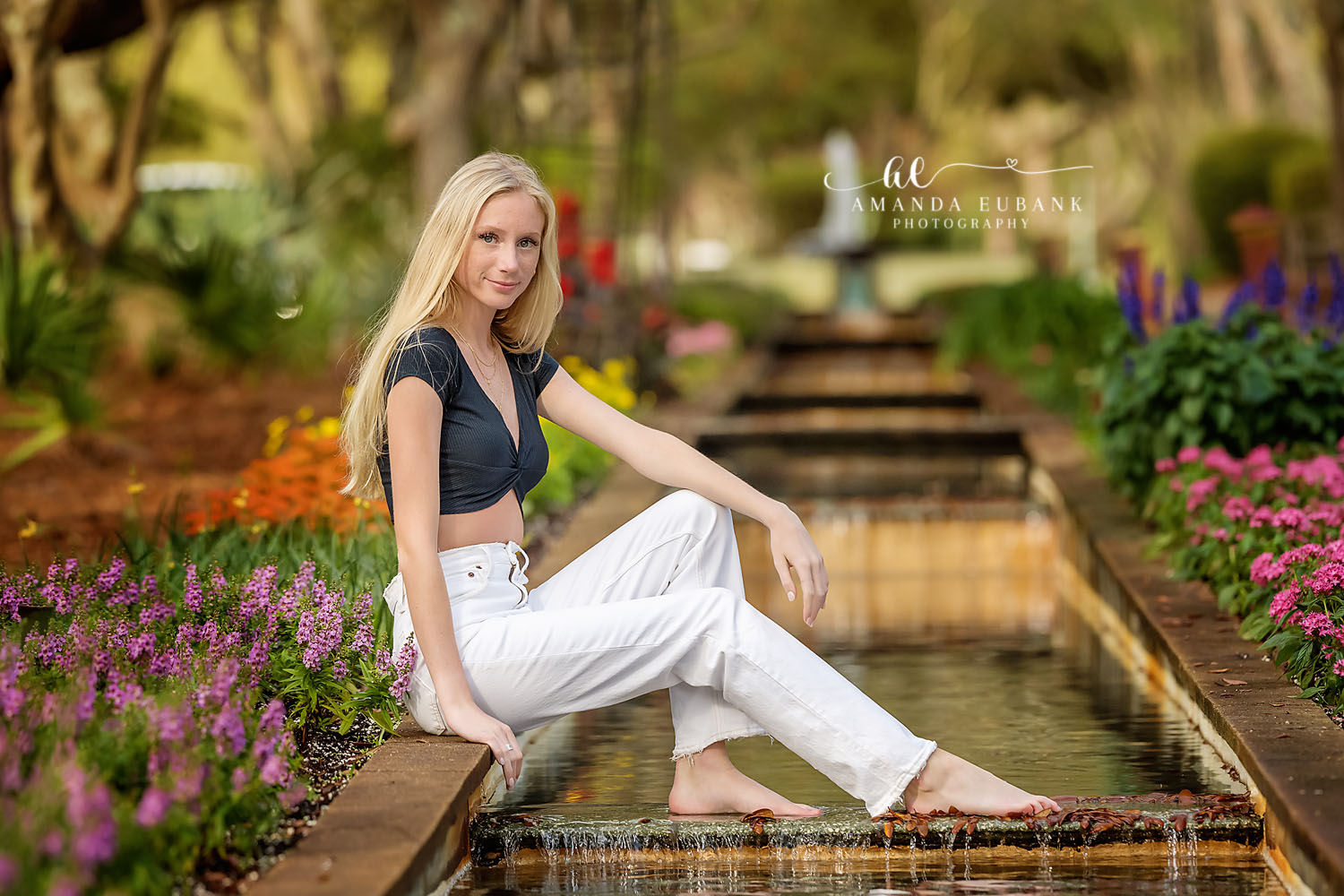Senior girl in a black top and white jeans seated by a garden fountain surrounded by colorful flowers, photographed by a Santa Rosa Beach senior photographer