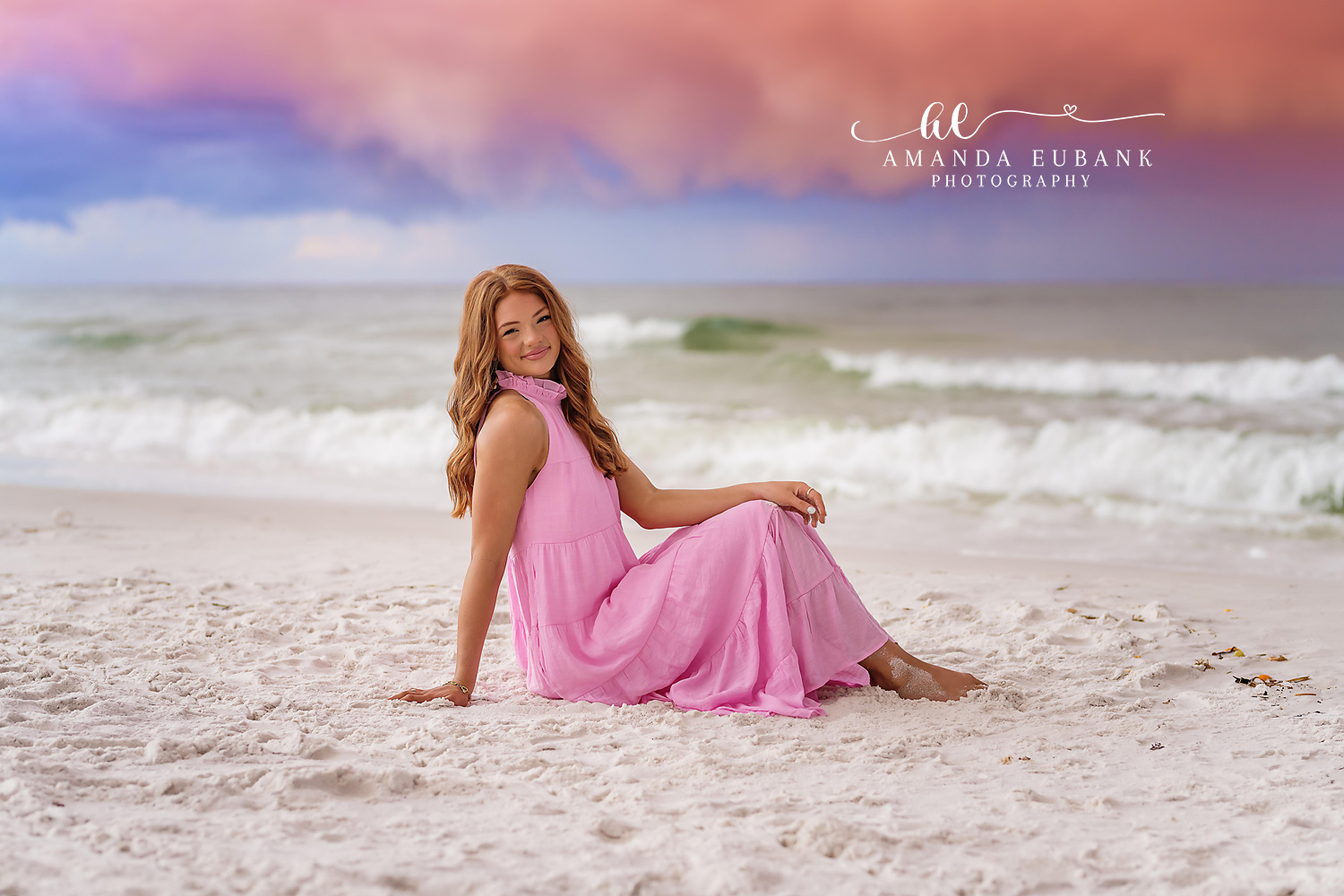 Senior girl in a long pink dress sitting on the white sand near the surf, photographed by a Santa Rosa Beach senior photographer