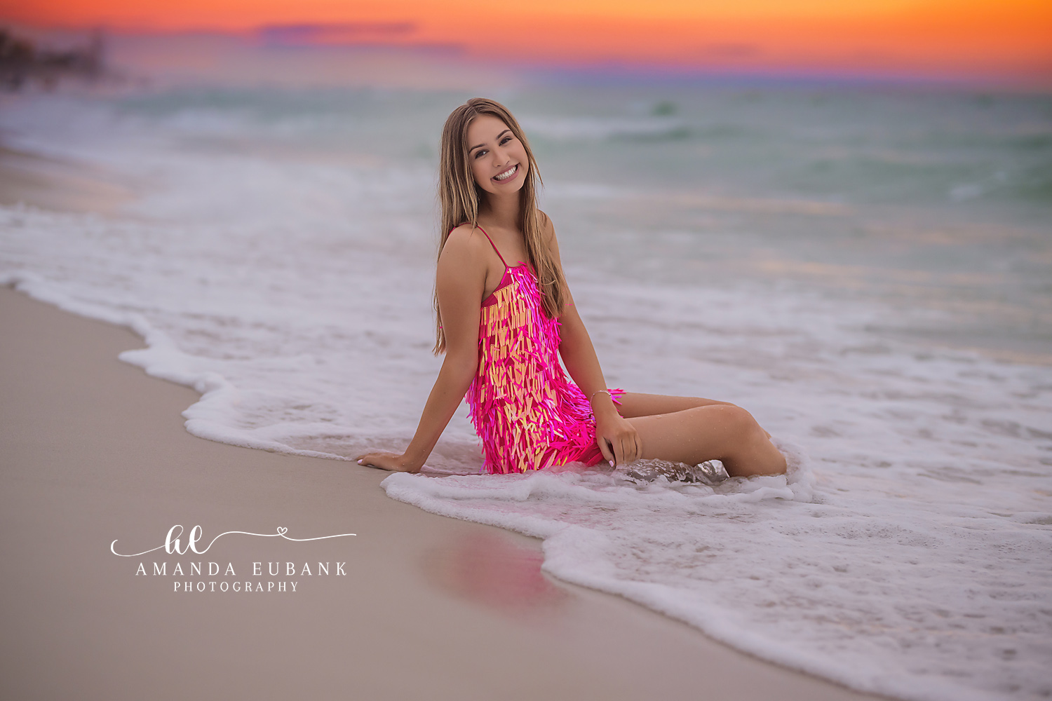 Senior girl in a pink and orange fringe dress sitting on the sand at the shoreline as the surf rolls in, photographed by a Santa Rosa Beach senior photographer