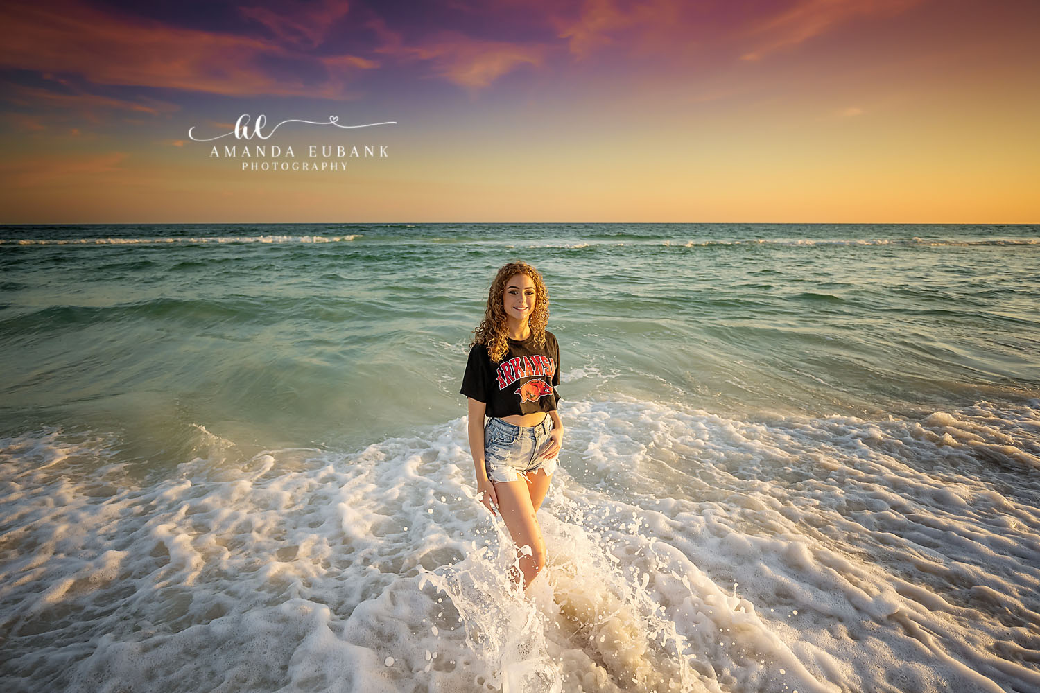 Senior girl in a college spirit tee and denim shorts standing in the breaking surf at golden hour, photographed by a Santa Rosa Beach senior photographer
