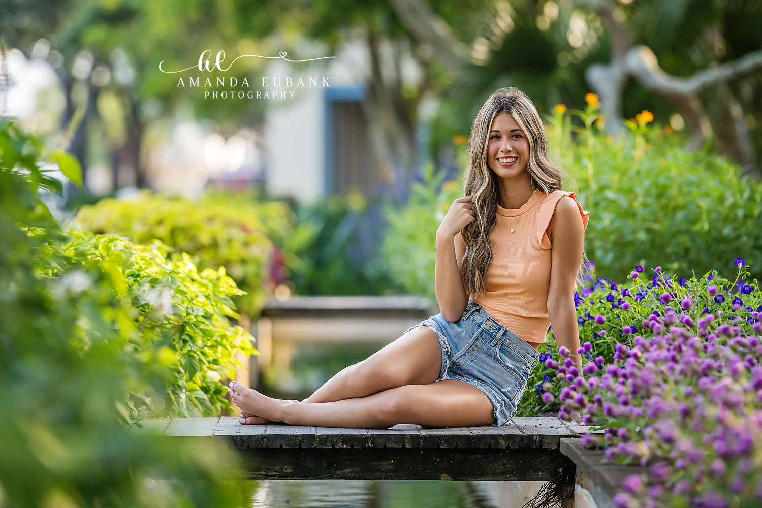 Senior girl in an orange top and denim shorts seated by a garden fountain surrounded by purple flowers, photographed by a Santa Rosa Beach senior photographer