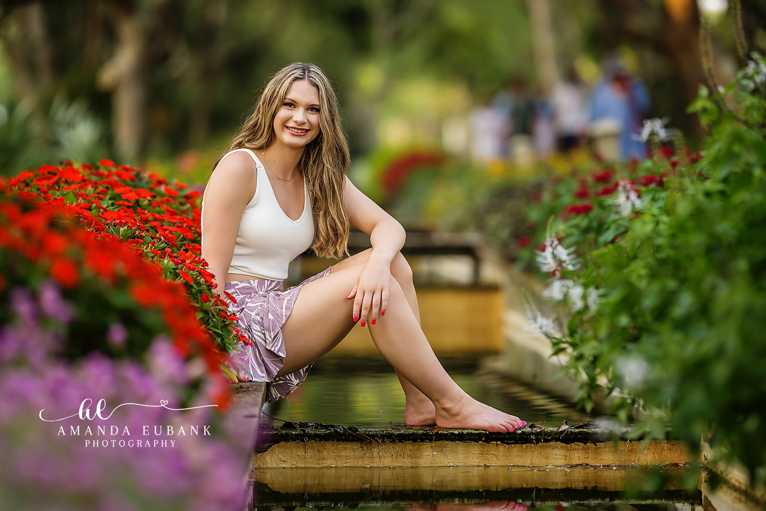 Senior girl in a white top and patterned shorts seated by a garden fountain surrounded by red and purple flowers, photographed by a Santa Rosa Beach senior photographer