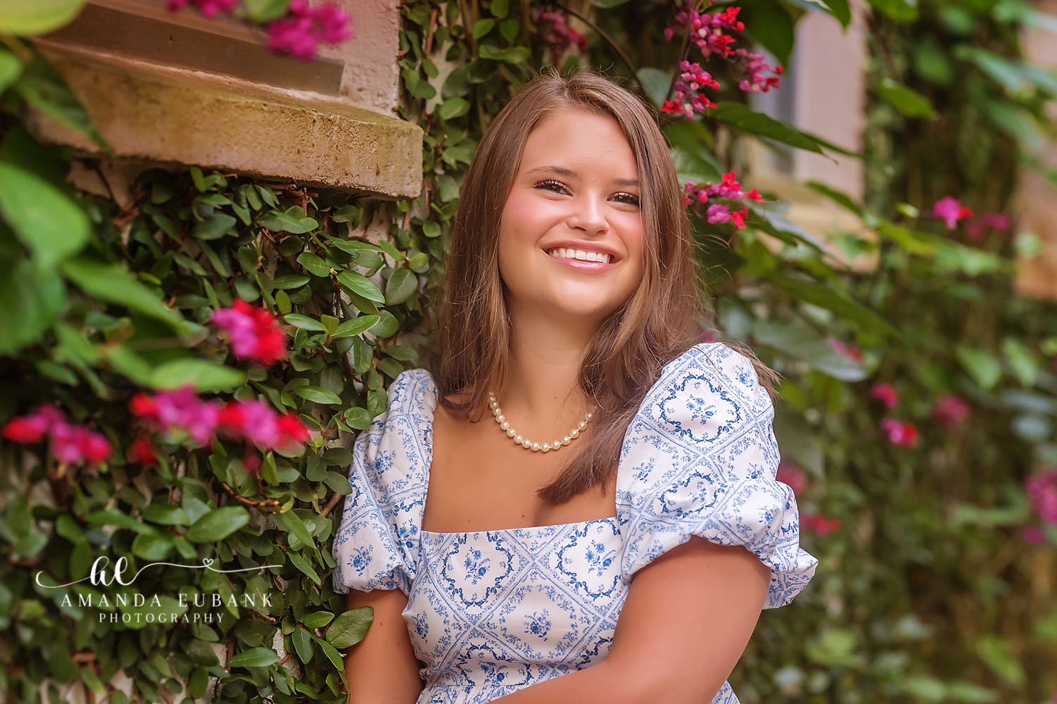 Senior girl in a blue and white patterned dress with a pearl necklace posed against a green hedge with pink bougainvillea, photographed by a Santa Rosa Beach senior photographer