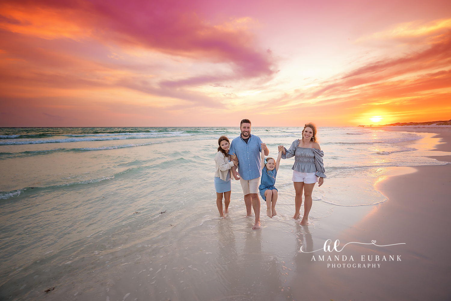 Family of four at the shoreline swinging their young daughter between them at a vivid orange sunset, photographed by a Santa Rosa Beach family photographer