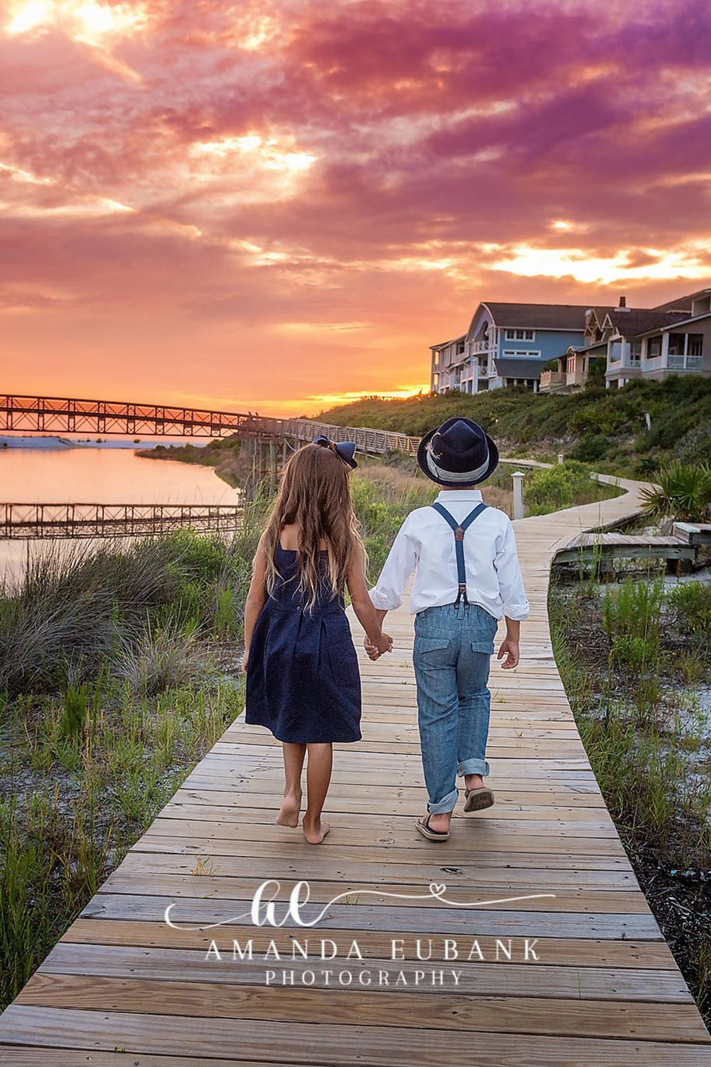 Santa Rosa Beach Family Photographer – Siblings Watching the Sunset Two young siblings seen from behind looking out over a coastal lagoon and boardwalk toward a vivid orange sunset, photographed by a Santa Rosa Beach family photographer