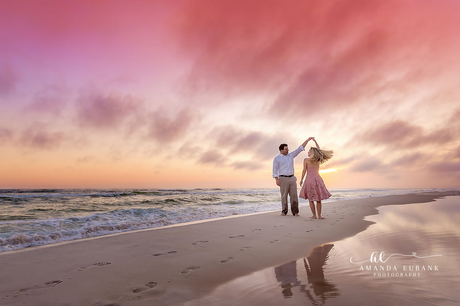 Man twirling a woman in a pink dress as they dance on the shoreline at a pastel sunset with reflections in the wet sand, photographed by a Santa Rosa Beach engagement photographer