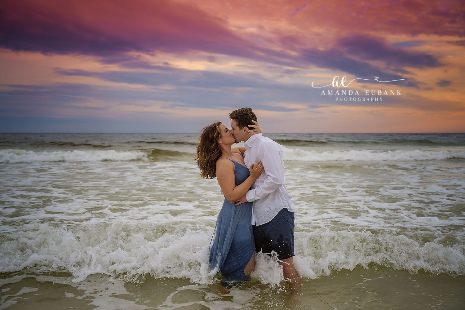 Couple kissing in the crashing surf at sunset, the woman in a blue dress, photographed by a Santa Rosa Beach engagement photographer