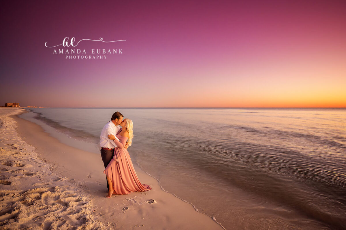 Couple kissing at the water’s edge against a vivid pink and orange sunset, the woman in a long pink dress, photographed by a Santa Rosa Beach engagement photographer