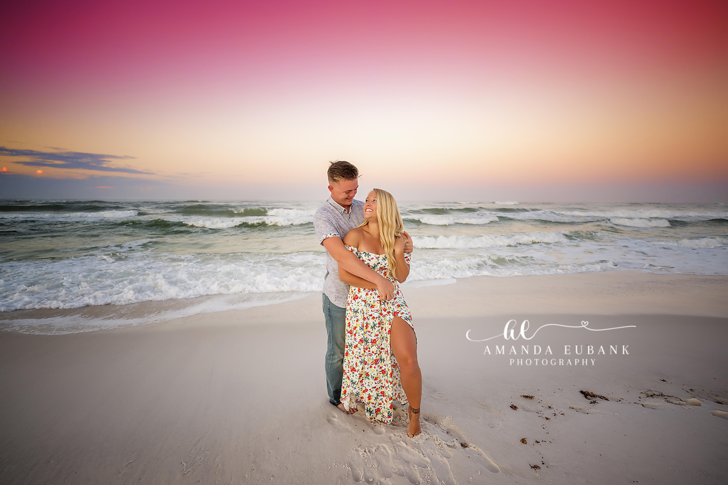 Couple embracing on the beach at sunset as the woman in a floral dress smiles up at the man, photographed by a Santa Rosa Beach engagement photographer