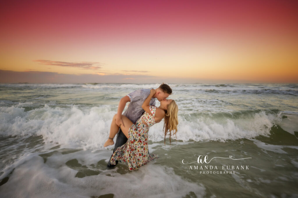 Man dipping a woman for a kiss amid the crashing waves at sunset, the woman in a floral dress, photographed by a Santa Rosa Beach engagement photographer