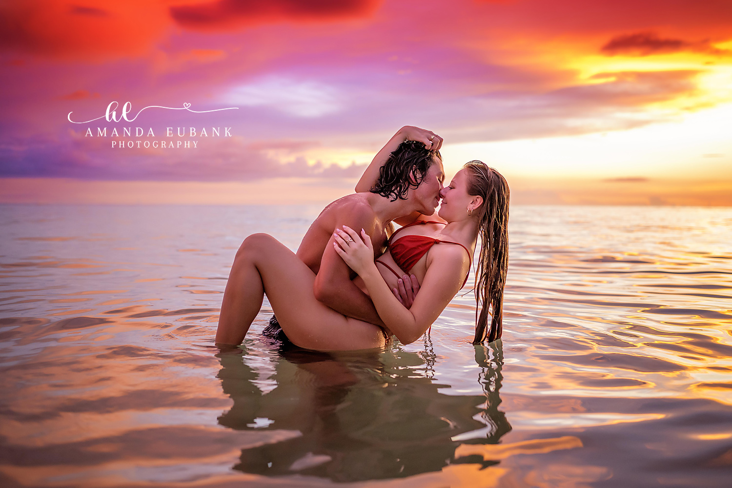 Santa Rosa Beach Engagement Photographer – Couple Embracing in the Shallows at Sunset Couple embracing closely while sitting together in the shallow water at sunset, the woman in a red swimsuit, photographed by a Santa Rosa Beach engagement photographer