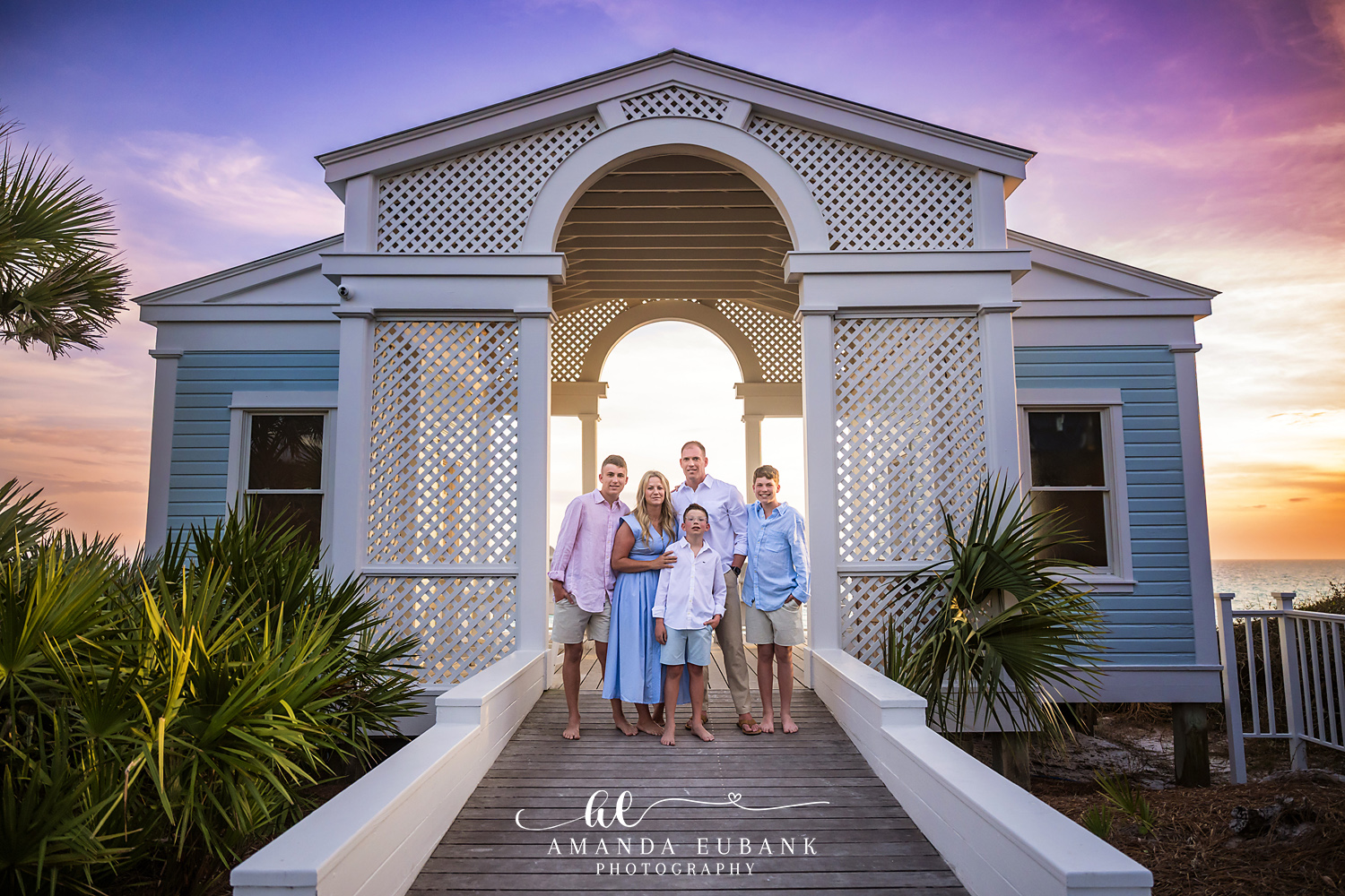 Santa Rosa Beach Family Photographer – Family at the Beach Cottage at Sunset Family of five posing on a coastal cottage walkway framed by a white lattice archway with the sunset over the ocean behind them, photographed by a Santa Rosa Beach family photographer