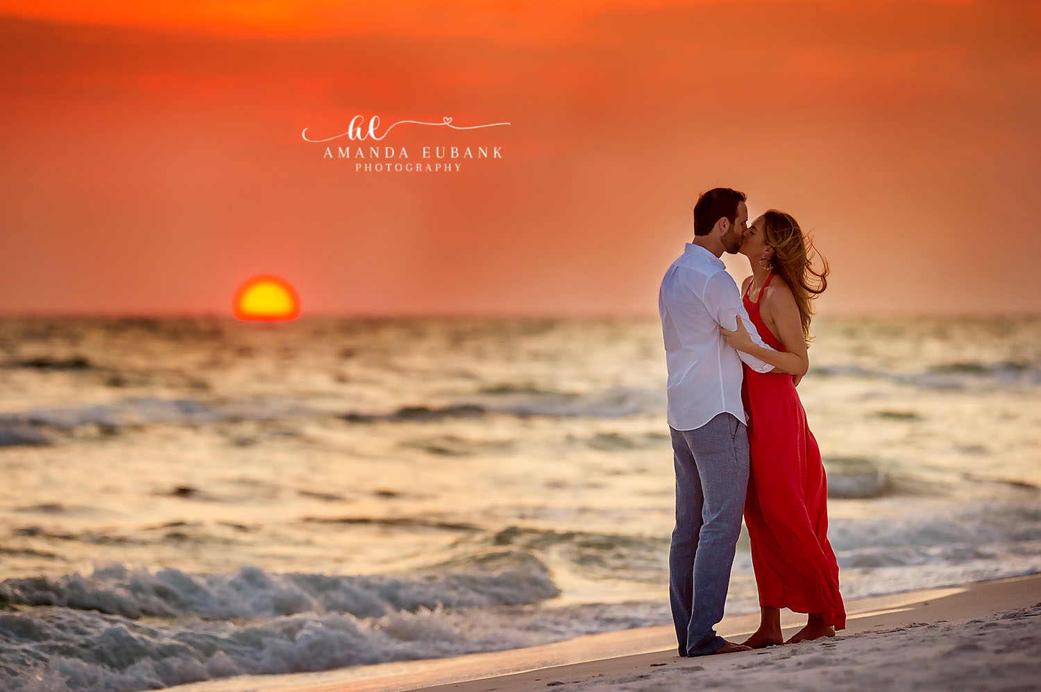 Couple kissing at the water’s edge as the sun sets on the horizon, the woman in a red dress, photographed by a Santa Rosa Beach engagement photographer