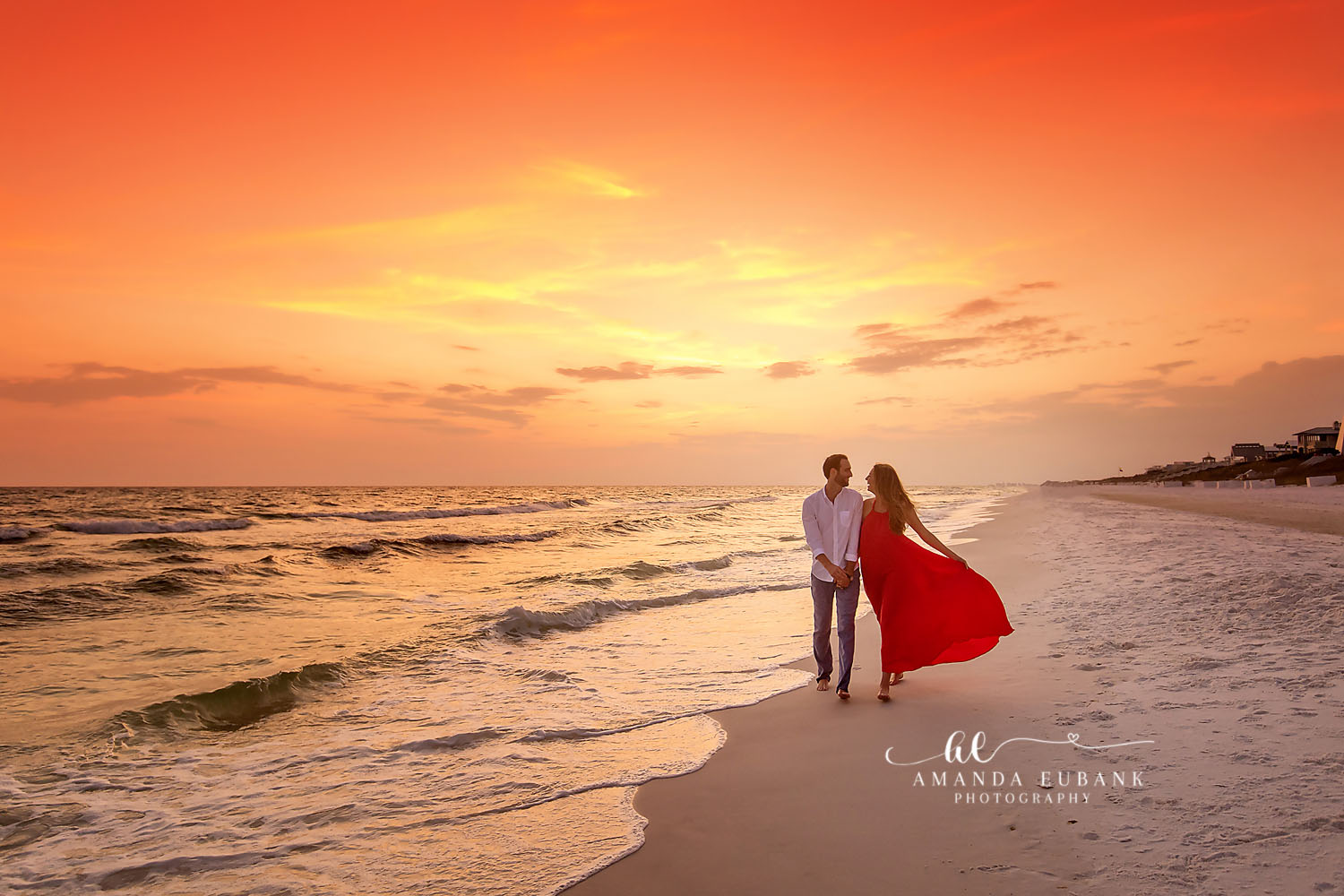 Couple walking together along the shoreline at golden sunset, the woman in a flowing red dress, photographed by a Santa Rosa Beach engagement photographer