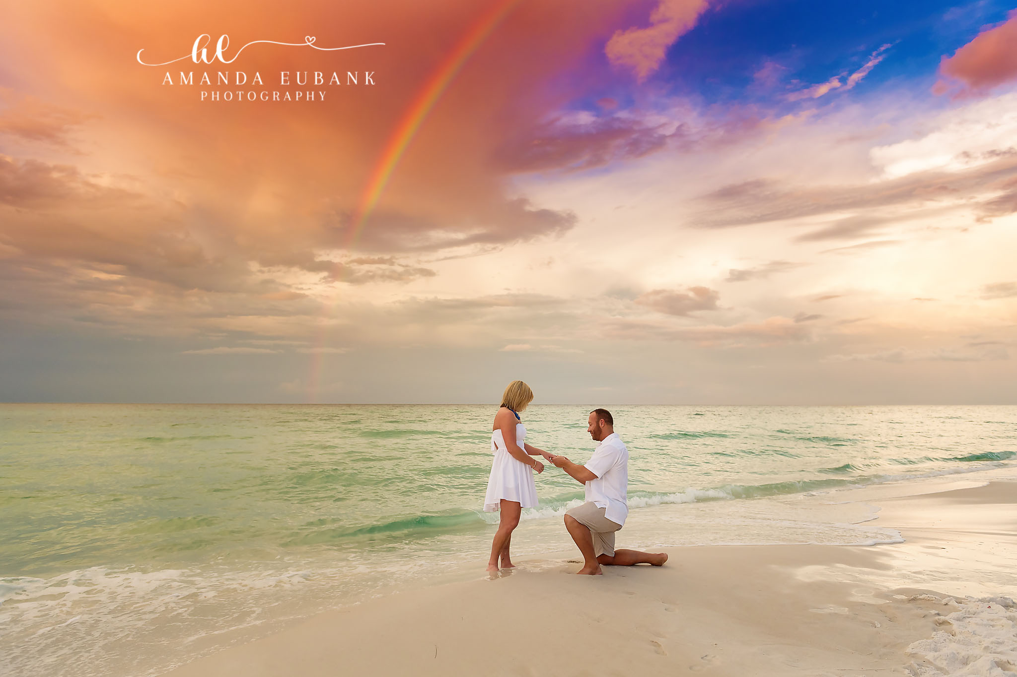 Santa Rosa Beach Engagement Photographer – Beach Proposal Beneath a Rainbow Man on one knee proposing to a woman in a white dress at the water’s edge with a faint rainbow in the sky, photographed by a Santa Rosa Beach engagement photographer