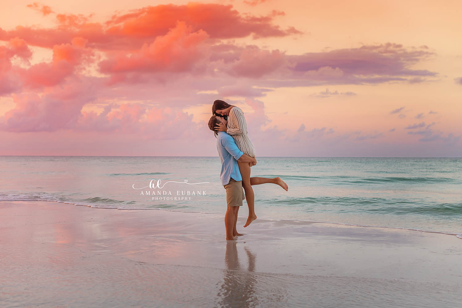 Man lifting a woman into his arms as they kiss on the shoreline at a pastel pink sunset, photographed by a Santa Rosa Beach engagement photographer