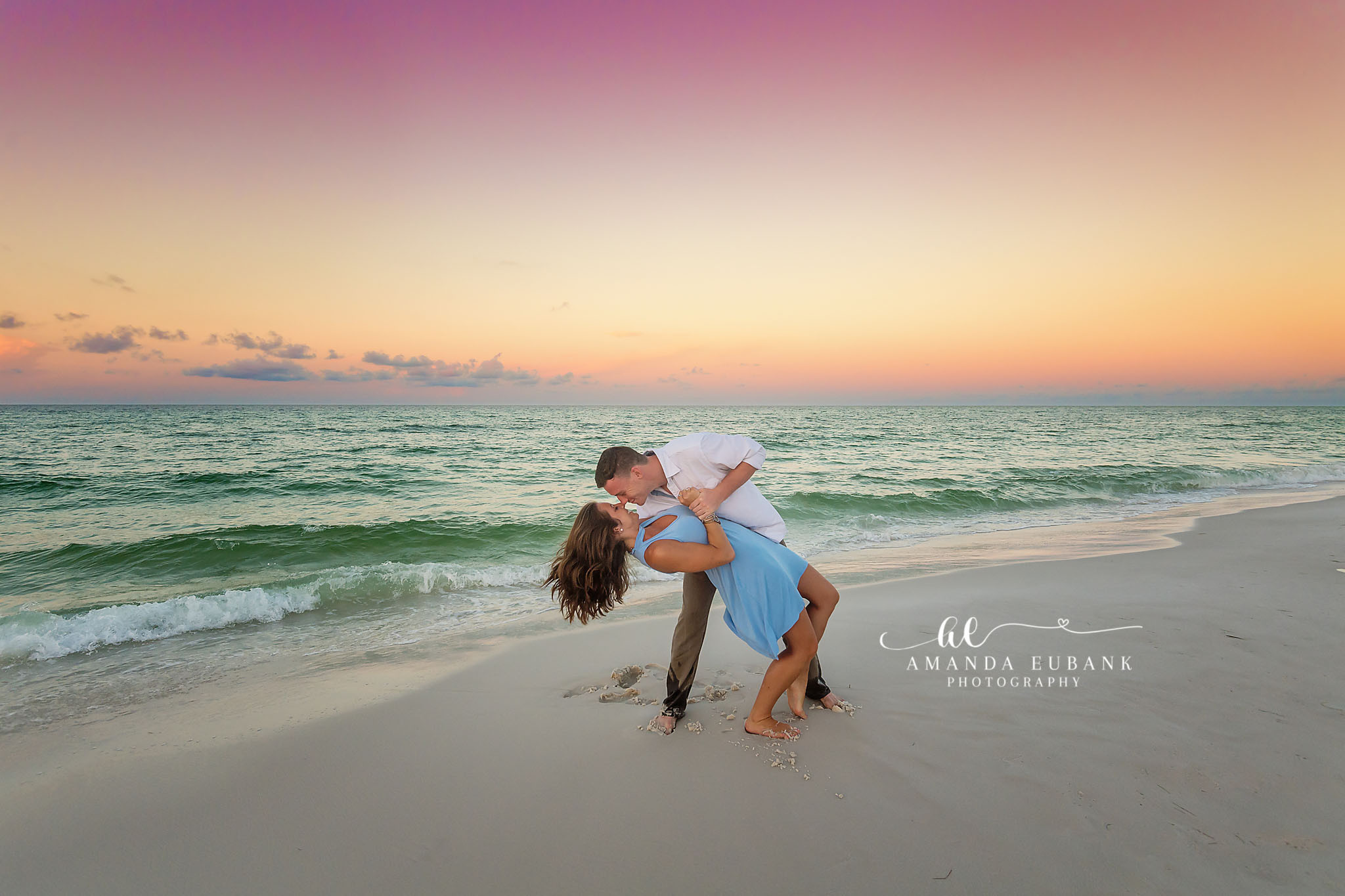 Man dipping a woman in a blue dress for a kiss on the shoreline at sunset, photographed by a Santa Rosa Beach engagement photographer
