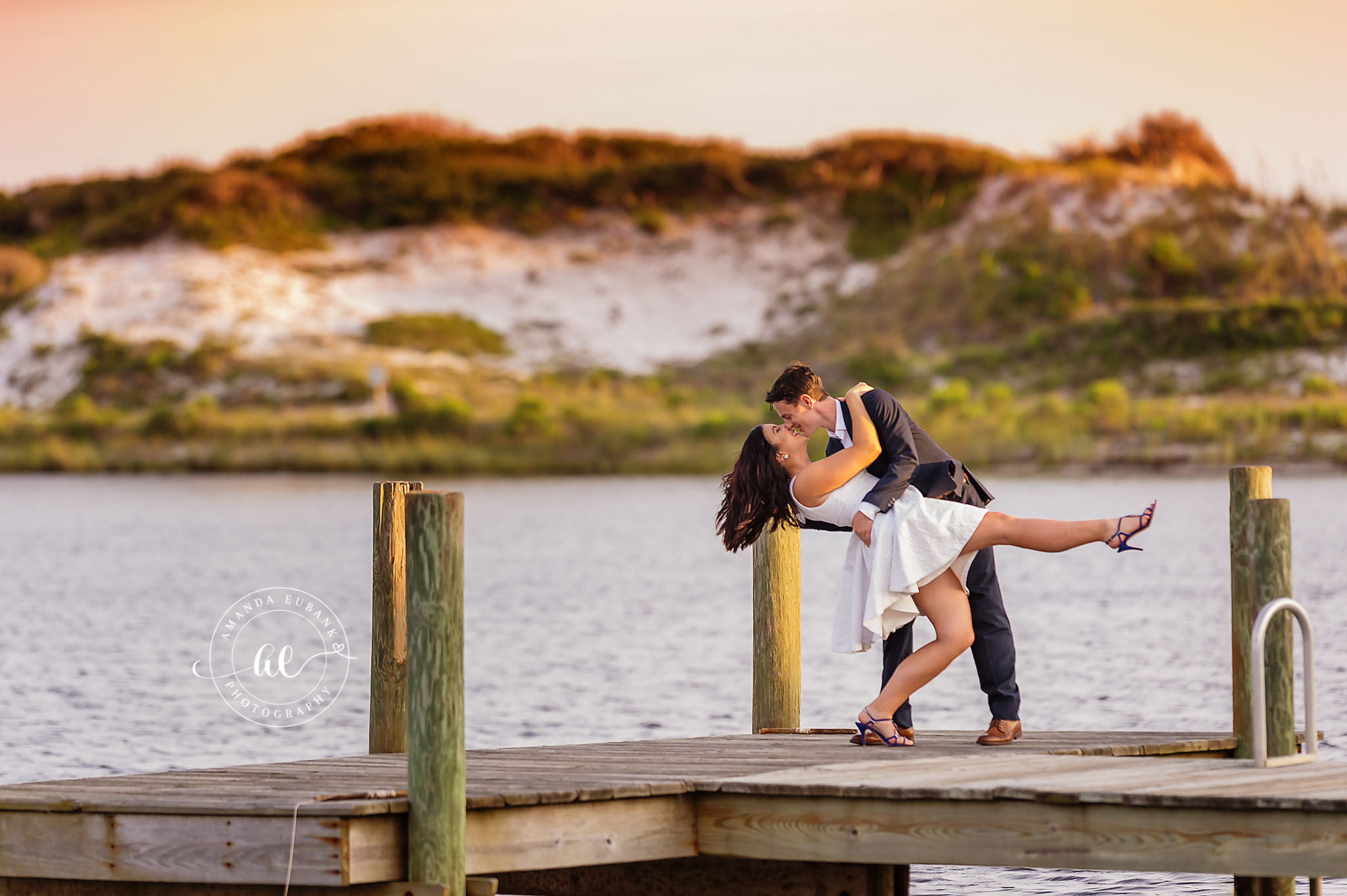 Man dipping a woman for a kiss on a wooden dock over the water, she in a white dress and he in a suit, photographed by a Santa Rosa Beach engagement photographer