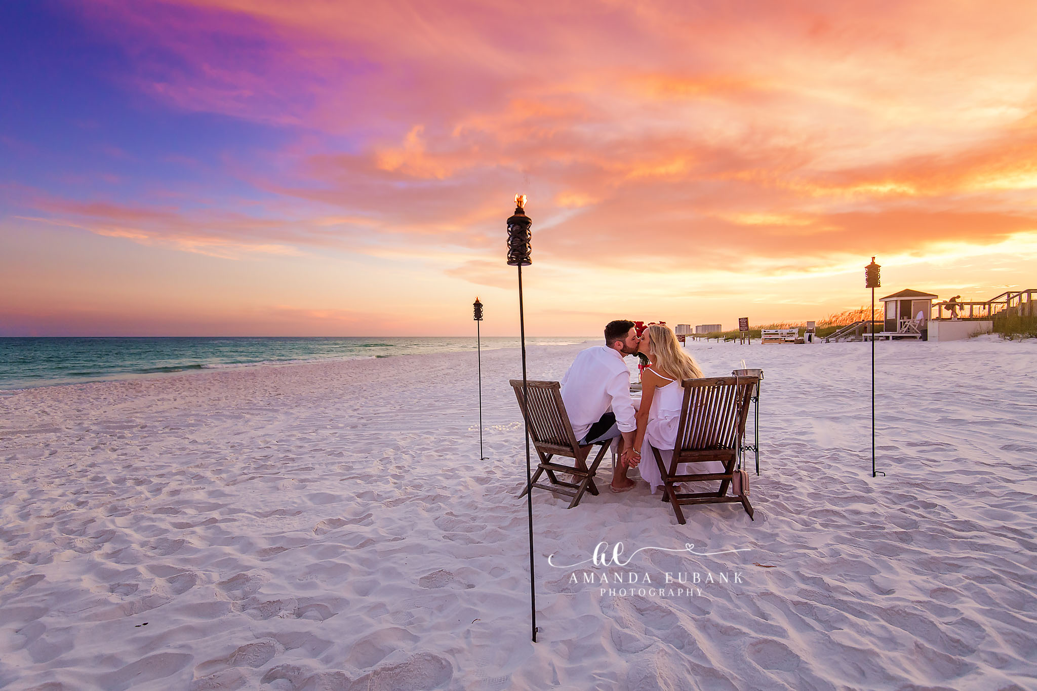 Couple kissing while seated in beach chairs between tiki torches at sunset on the white sand, photographed by a Santa Rosa Beach engagement photographer